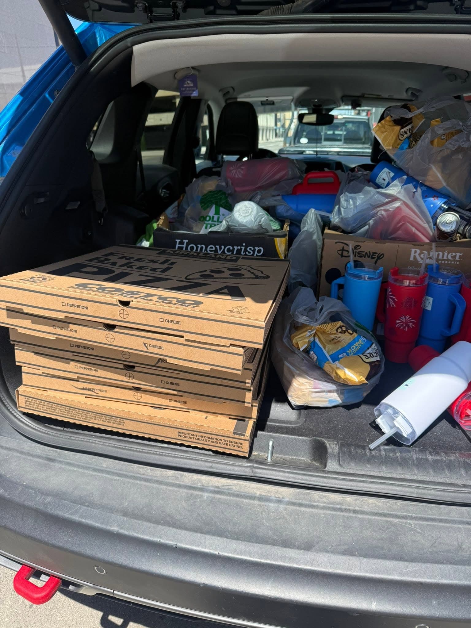 A car trunk loaded with pizza boxes, fresh apples, drink tumblers and bagged supplies for an outreach shift.