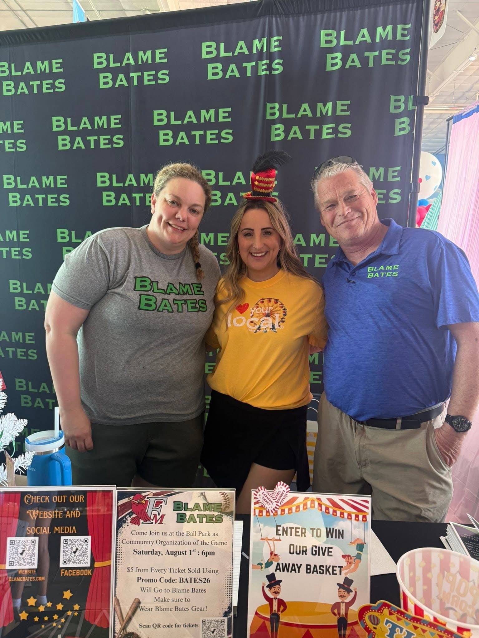 Three people at a Blame Bates table in front of a branded backdrop at a community event.