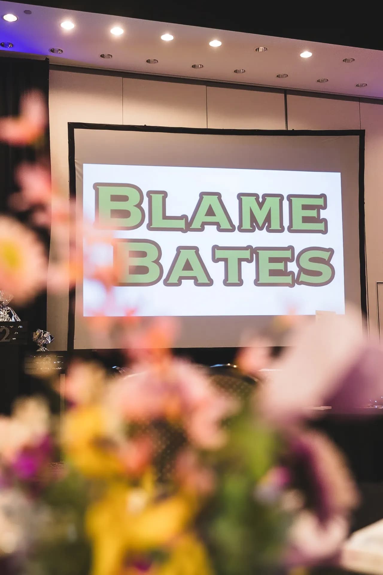 The Blame Bates logo on a projection screen behind a floral centerpiece.