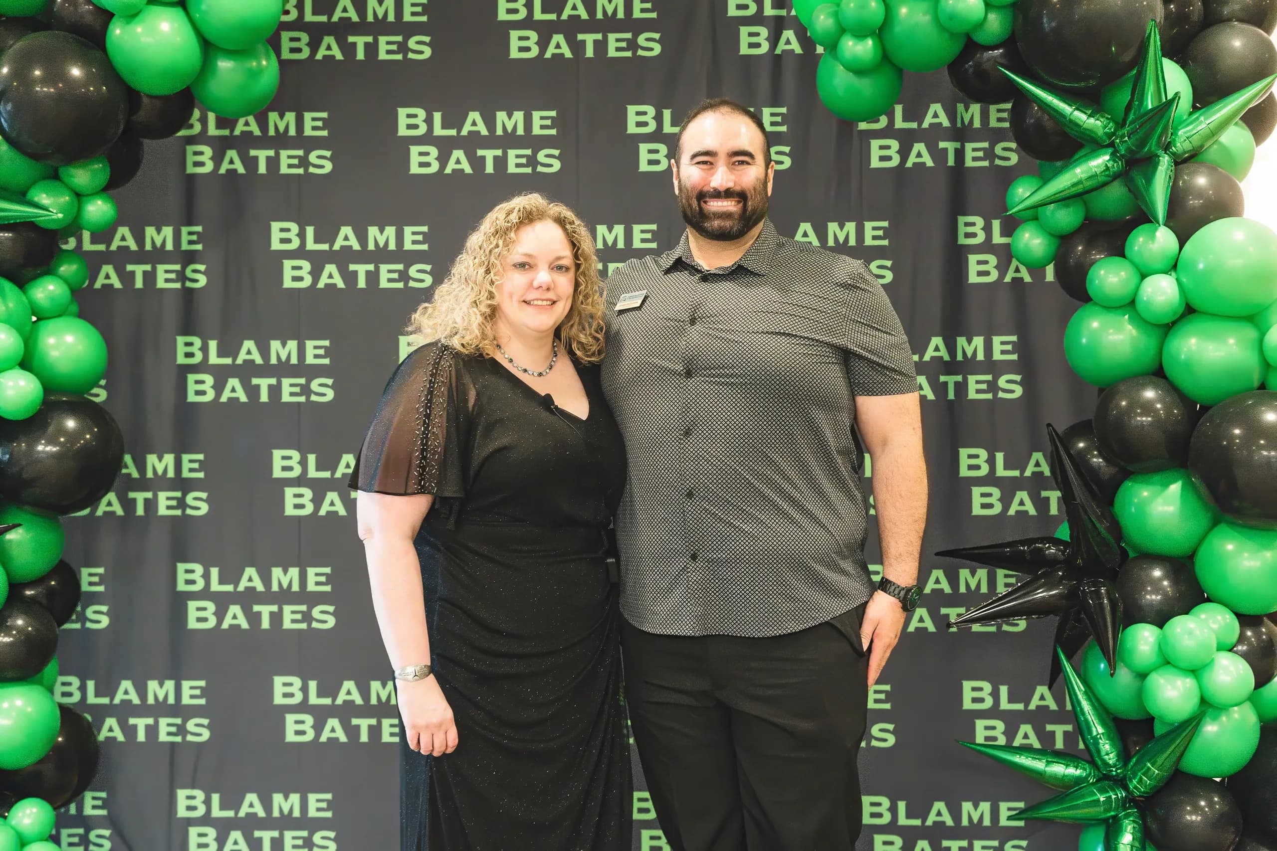 Two guests photographed in front of a green and black Blame Bates balloon wall.