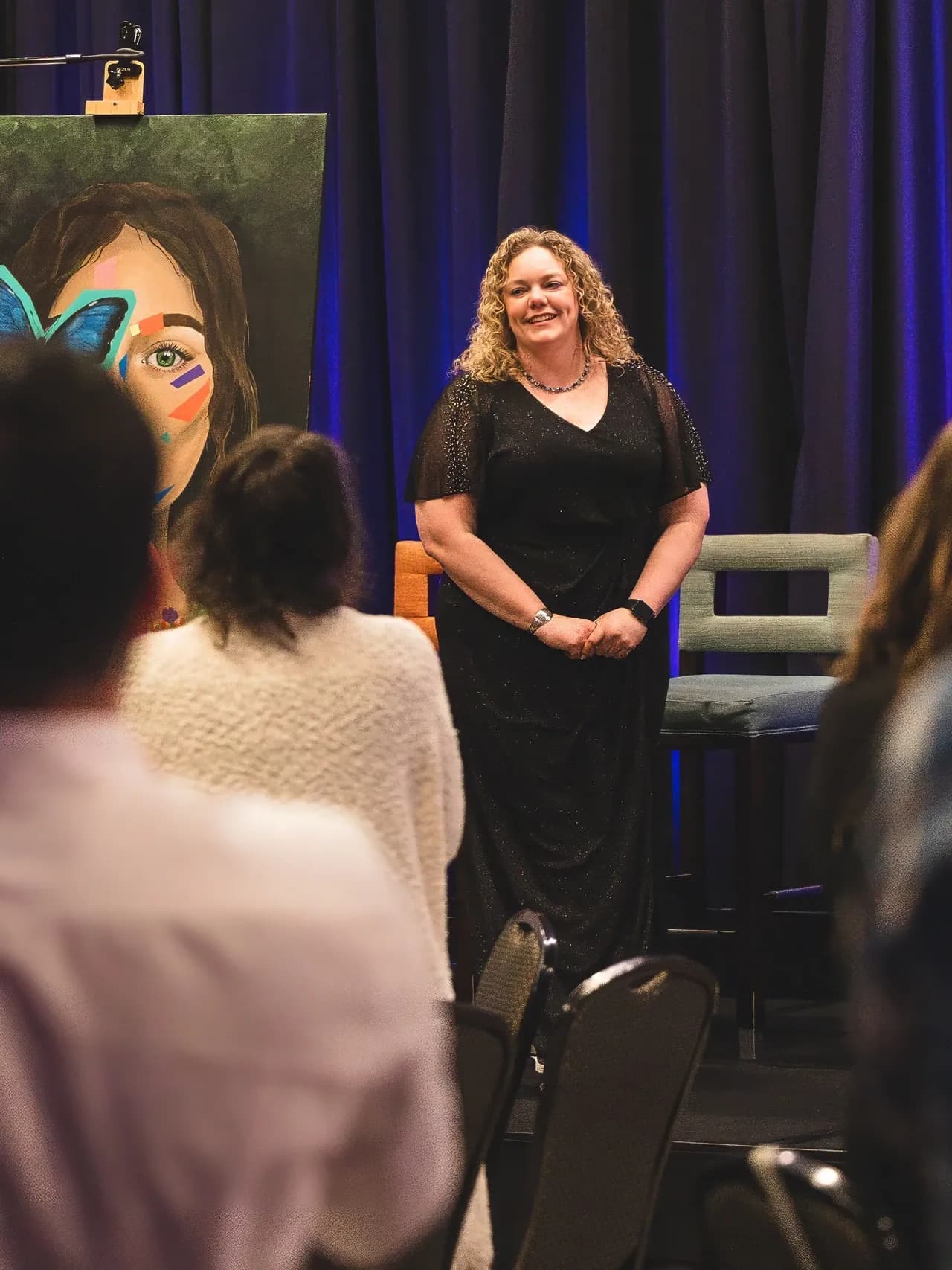 Blame Bates founder Ashley Bates speaking to seated guests at the Paint the Night Away gala, beside a painted portrait on an easel.
