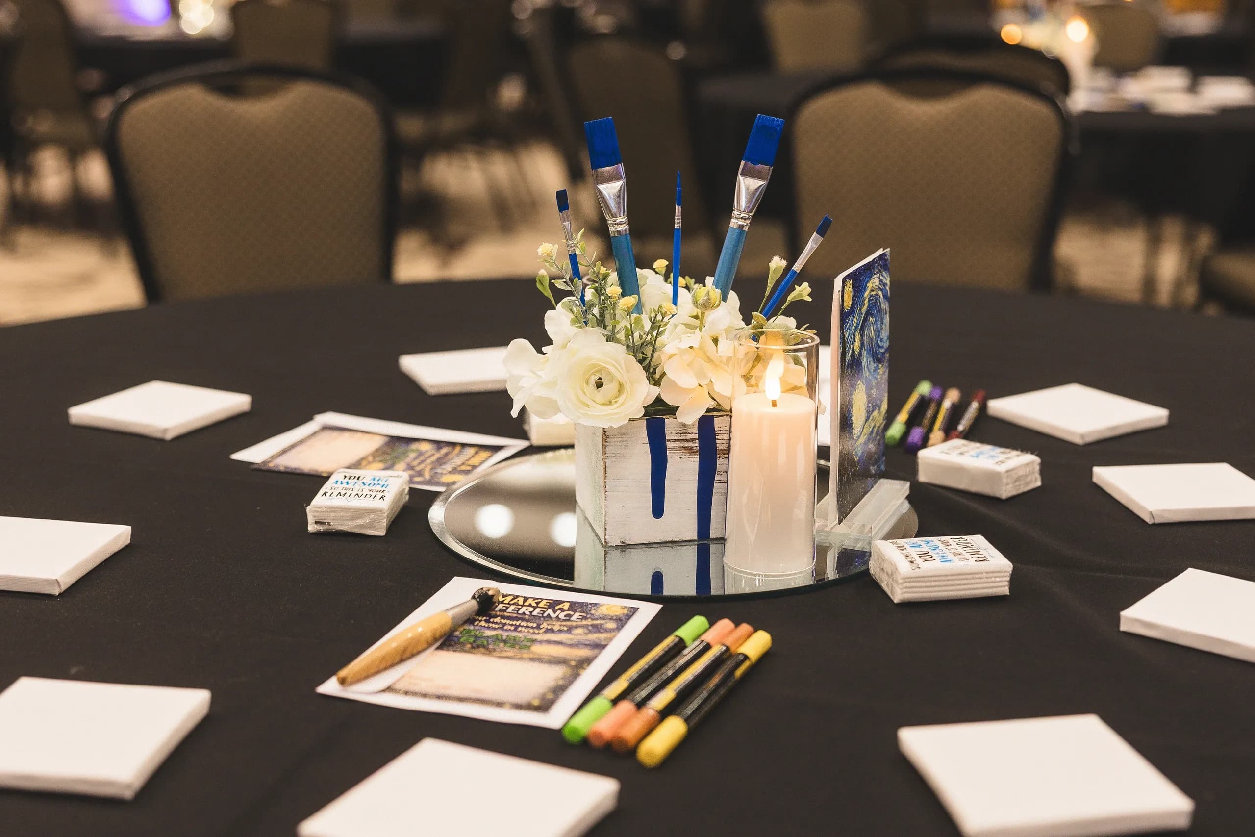 A banquet table set with paintbrushes, paints and a candle for the Paint the Night Away theme.