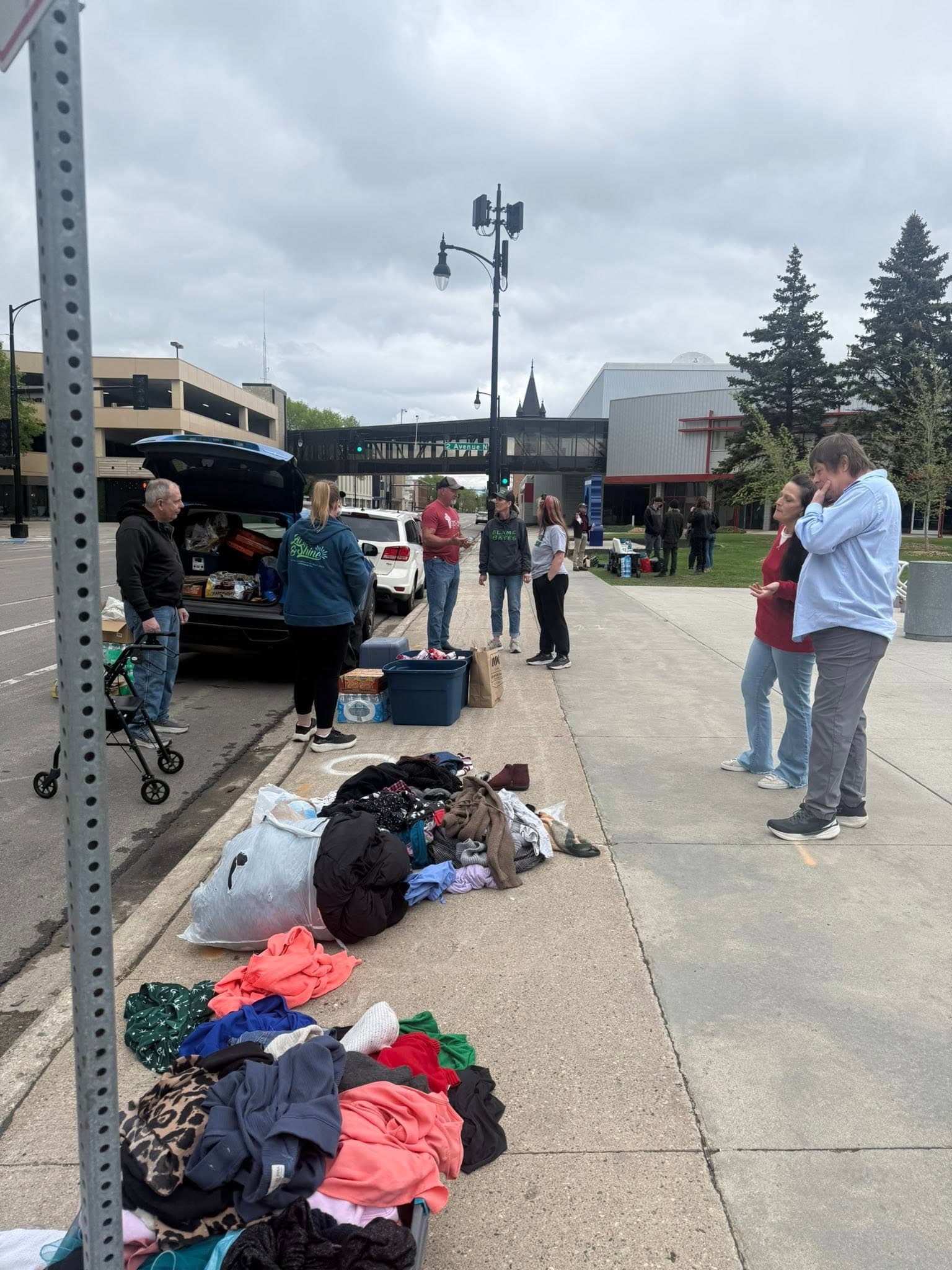 People gathered on a downtown Fargo sidewalk beside an open car trunk, with clothing and supplies laid out on the pavement.
