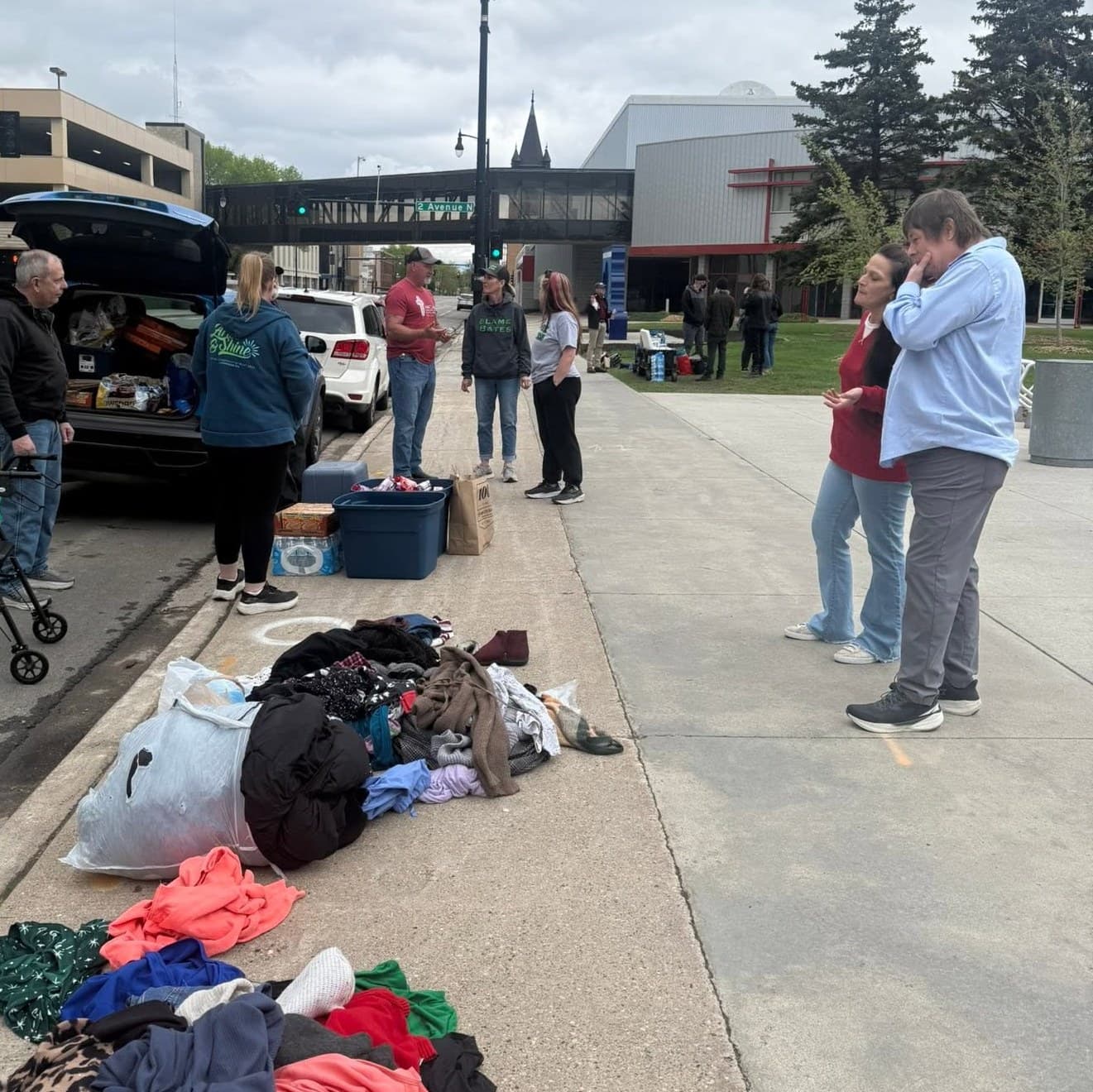 Volunteers hand out food and clothing from an open car trunk on a downtown Fargo sidewalk. Supplies are laid out along the pavement and two people talk at the edge of the group.